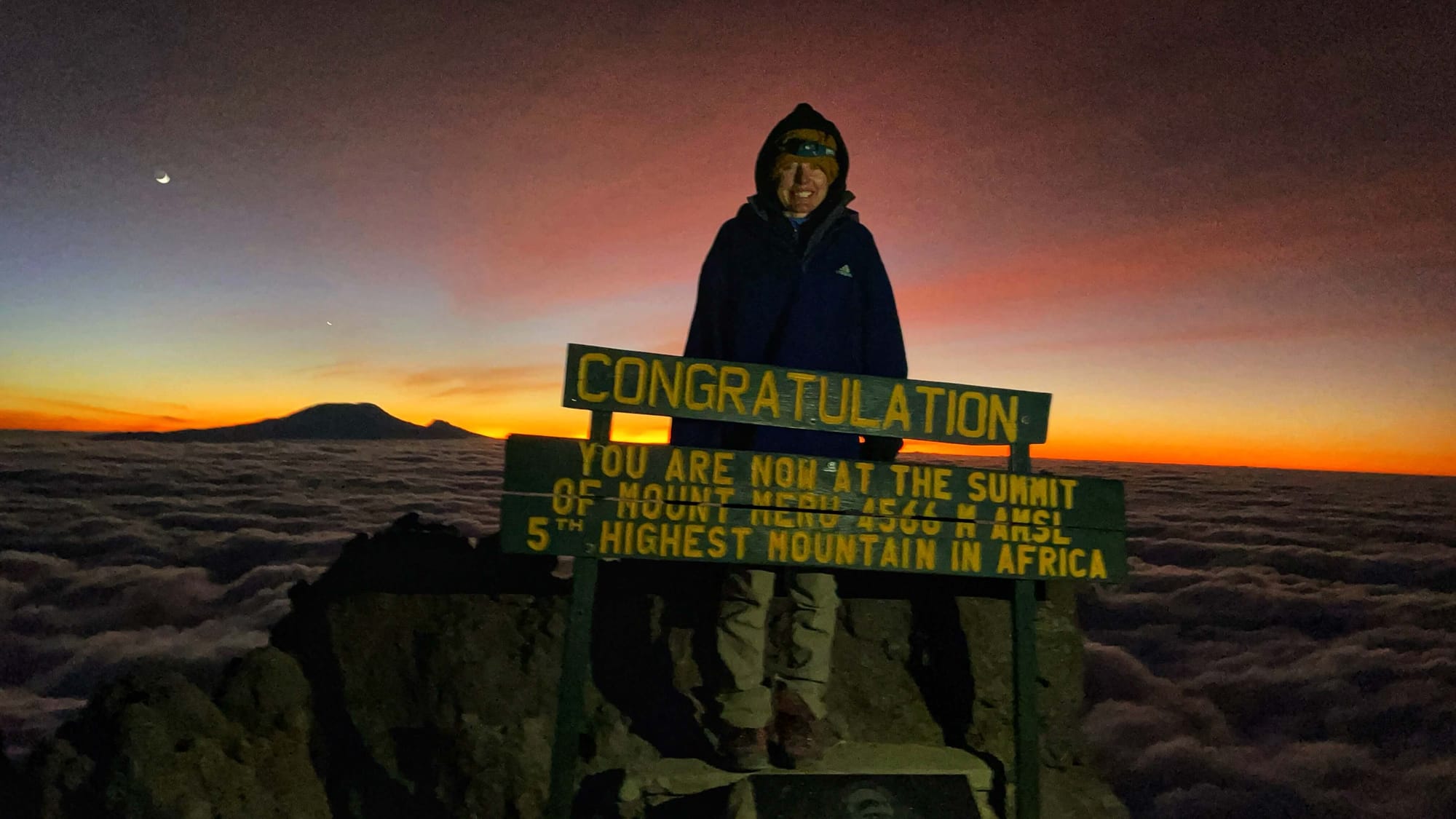 In the dark night sky with an orange glow on the horizon, Clara Ritger poses at the peak of Mount Meru with Mount Kilimanjaro in the background. A sign reads: "Congratulation you are now at the summit of Mount Meru 4566 M AMSL 5th highest mountain in Africa."