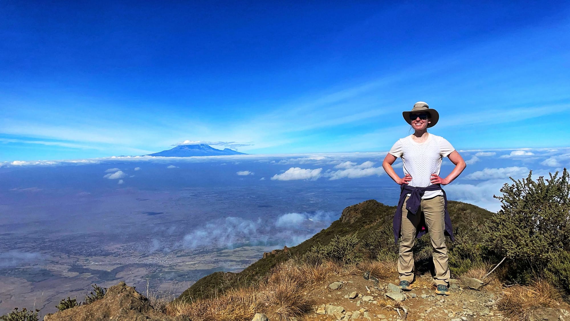 Clara Ritger with her hands on her hips and a large mountain in the background to her left. The sky is clear, and deep blue.