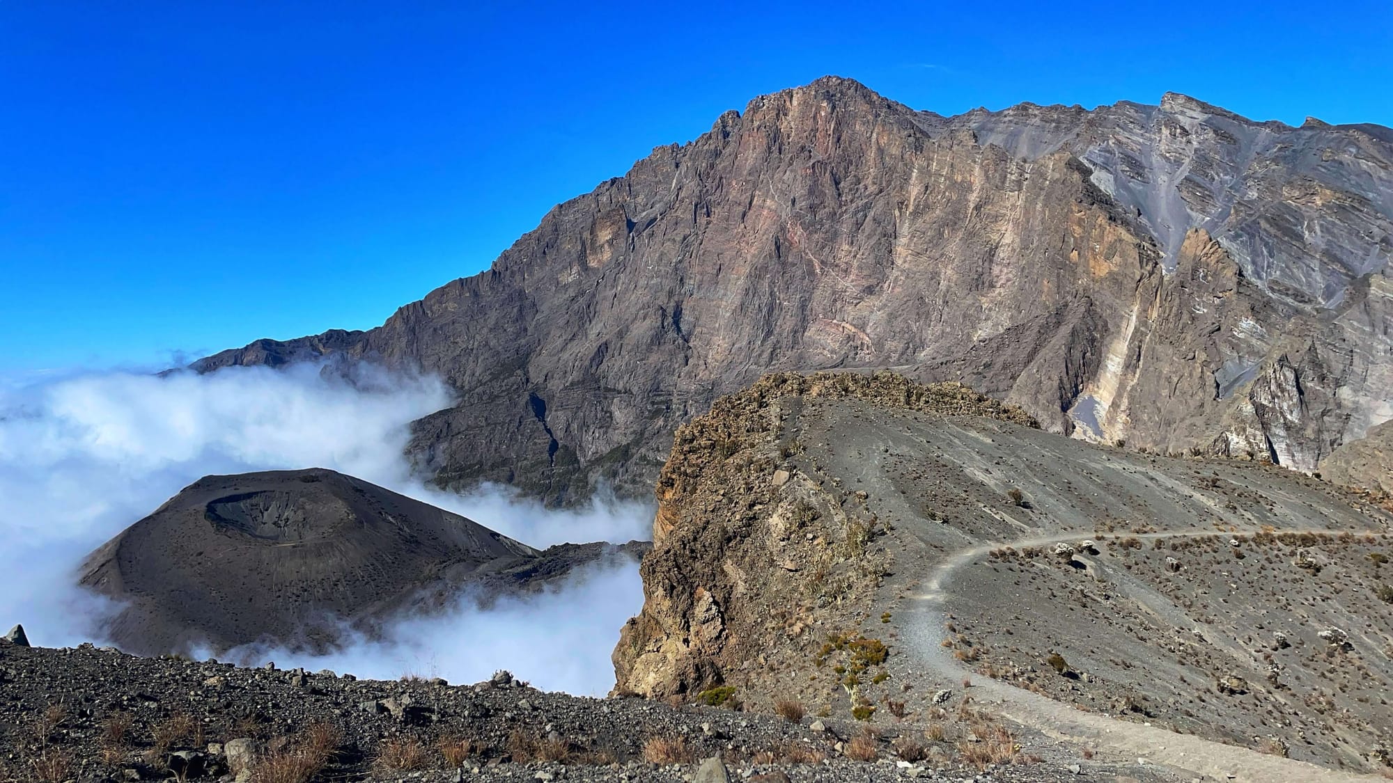 Clouds surround a cinder cone, a black sandy conical formation with a circular indent like a thumbprint. A jagged rocky peak rises up high above it, with a thin trail curving around the ridge line.