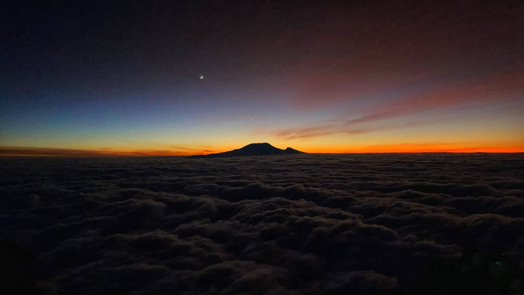 A crescent moon in a blue and deep purple night sky. A mountain with a large peak and a smaller peak like a knob sits above dark clouds. The horizon is orange and yellow, just before sunrise.