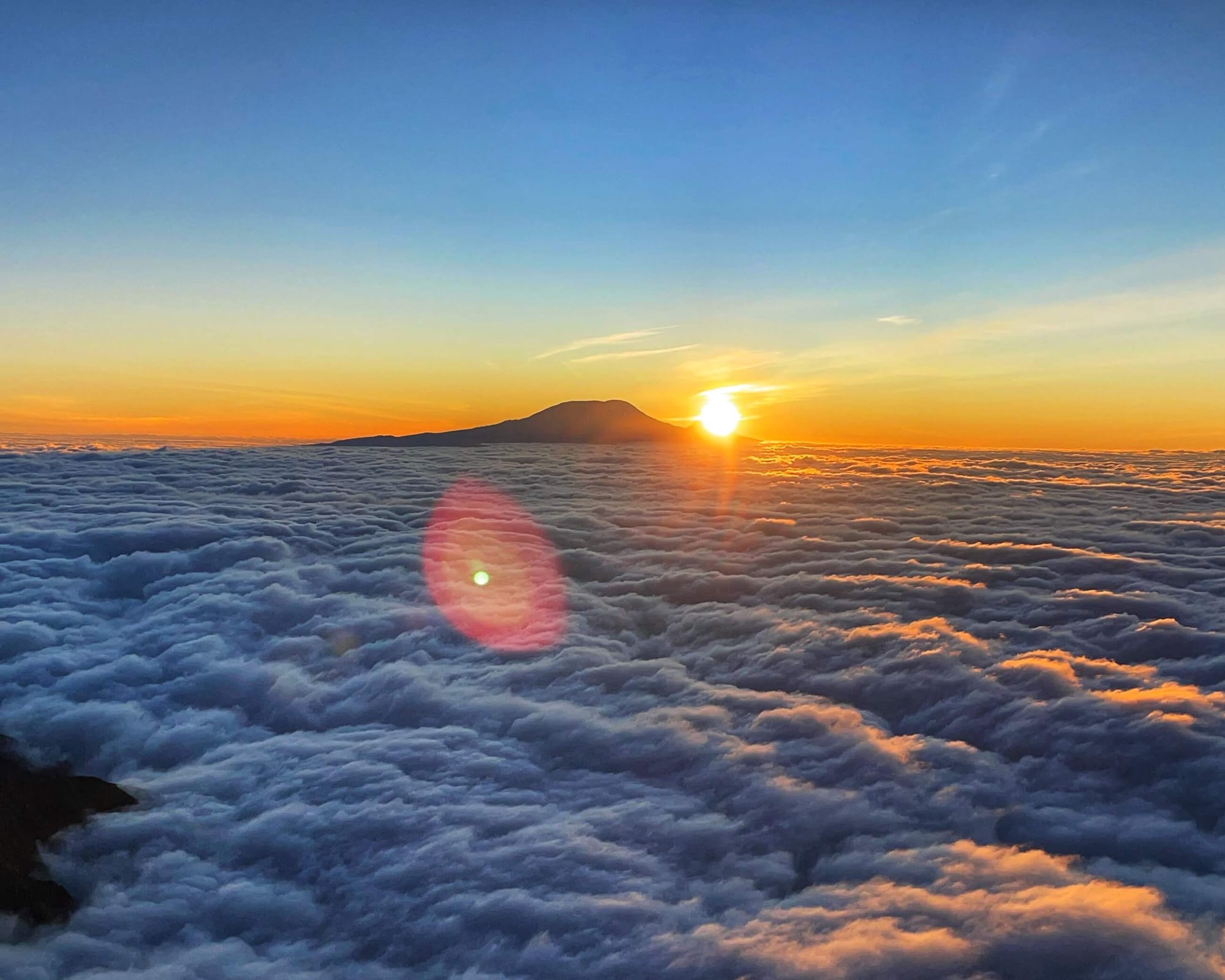 The sun rises over the corner of Mawenzi Peak on Mount Kilimanjaro, casting a glow on the lens. Clouds are painted orange. The sky is a deep blue, and the horizon a mix of orange and yellow.