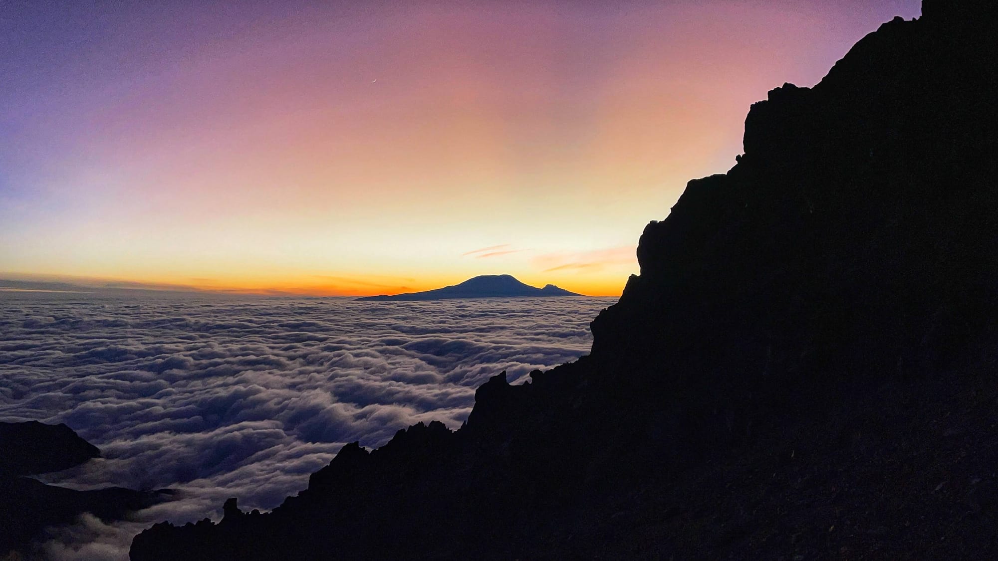 A large mountain is seen above the clouds, in a purple, pink, yellow and orange sky, as night turns to day.
