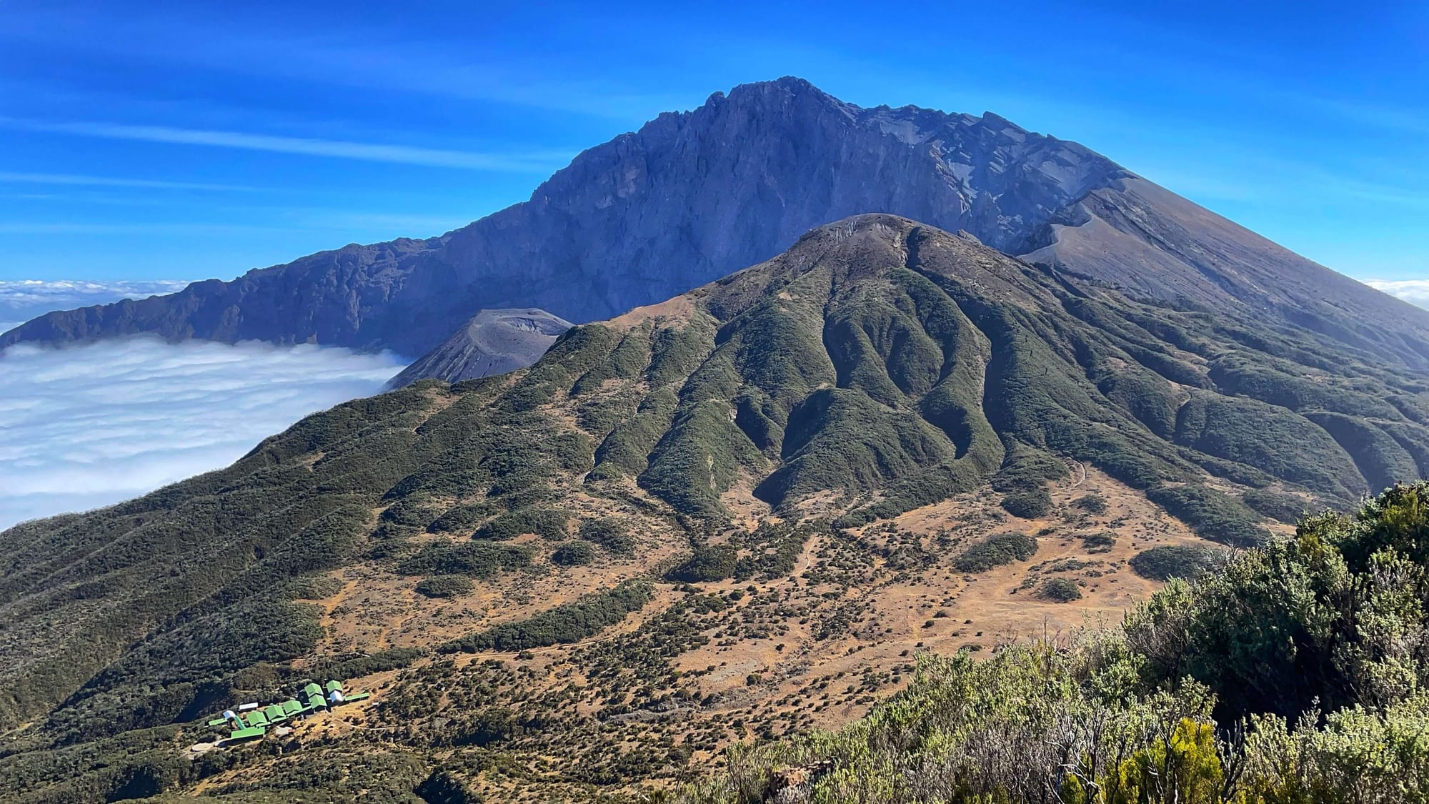 A half-moon shaped volcano with a crinkly ridgeline and a dark grey cider cone peeking out. Green roofed huts are seen below.