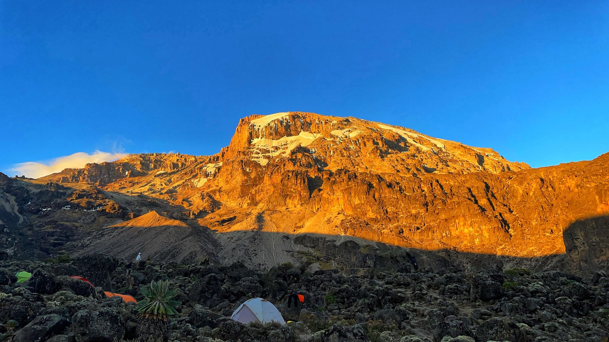 Orange sunset light hits a tall mountain peak. A few tents can be seen below.