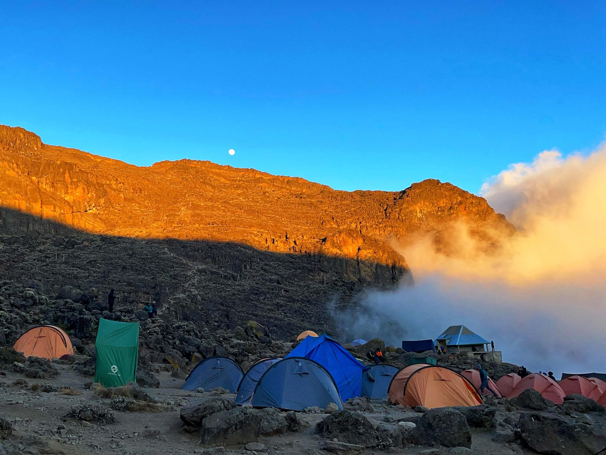 Colorful tents are shaded by the mountain as sunset casts a deep orange glow and the moon rises above the crest.