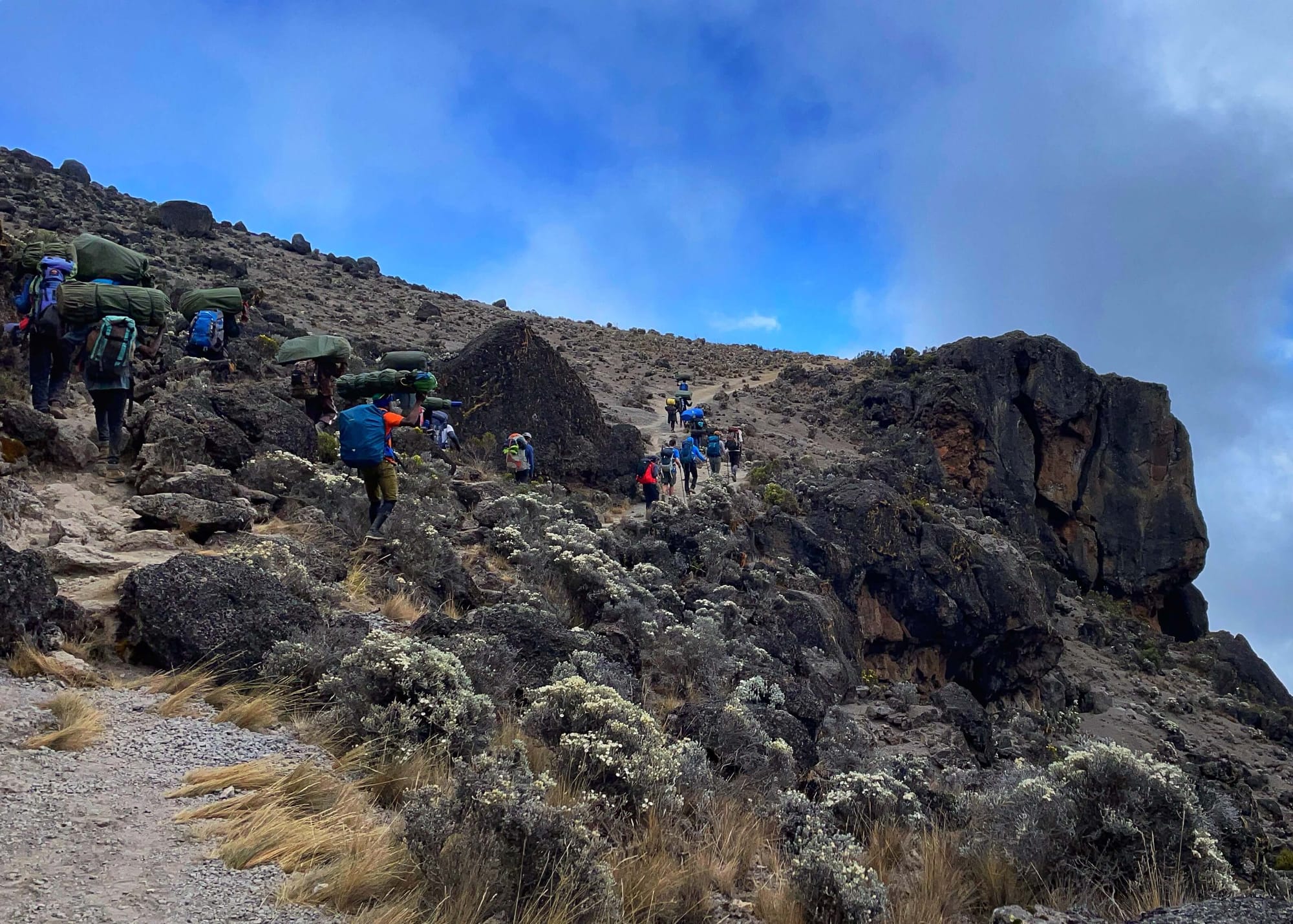 People with big green bags balanced on their heads trail behind hikers with poles.