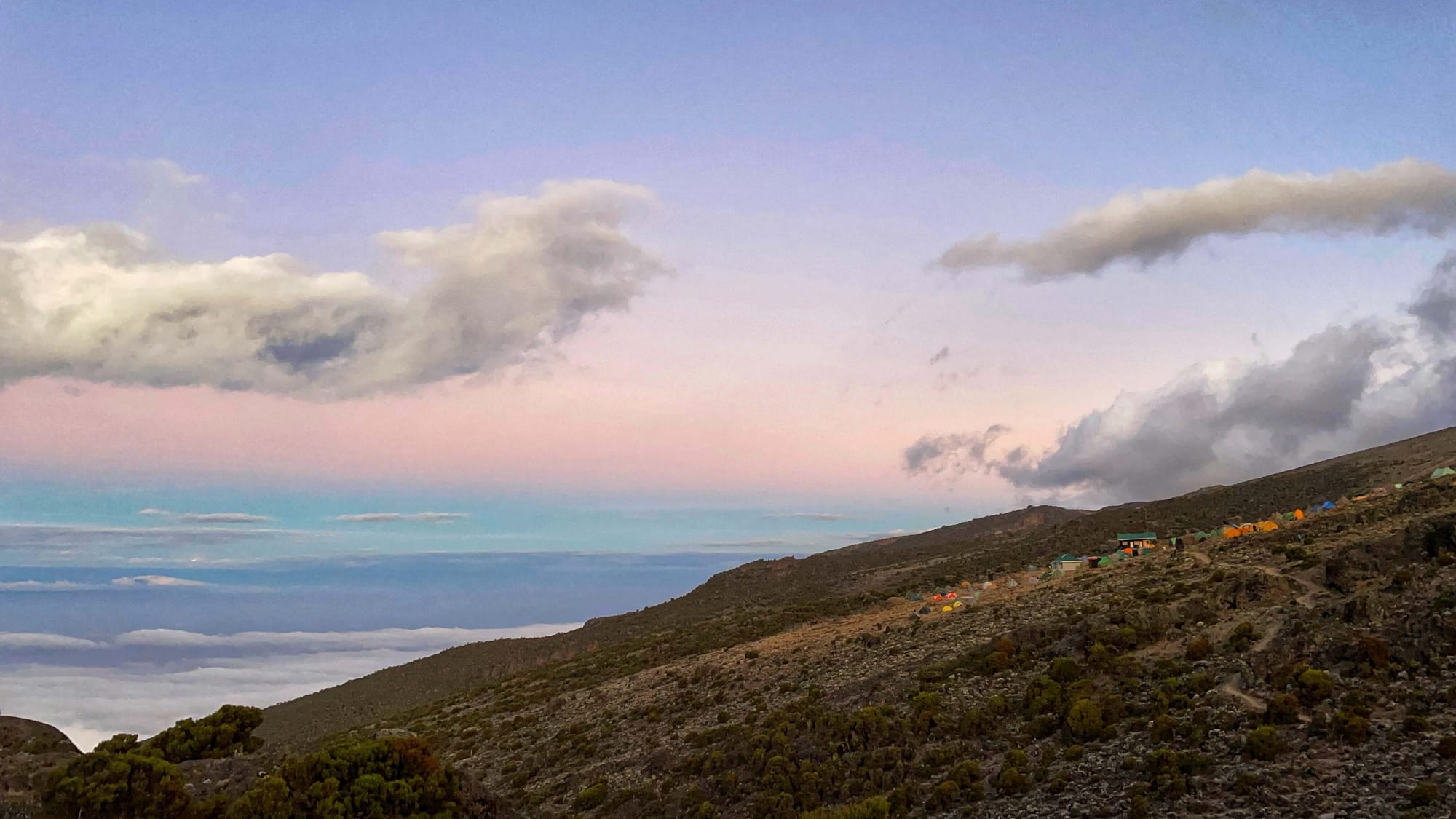 Colorful tents dot the side of a mountain at sunrise. The sky is shades of pink, purple, and blue pastel colors.