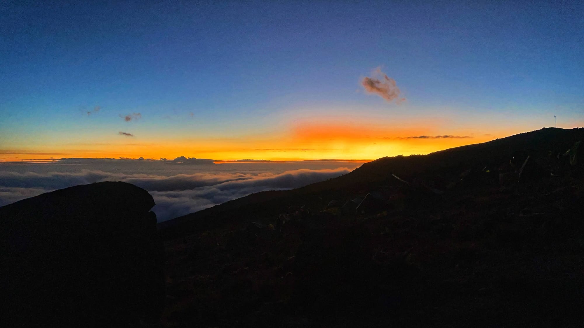 A deep blue sky and a horizon of bright yellow and orange. The foreground is in shadow, but the clouds below signal the side of a mountain.