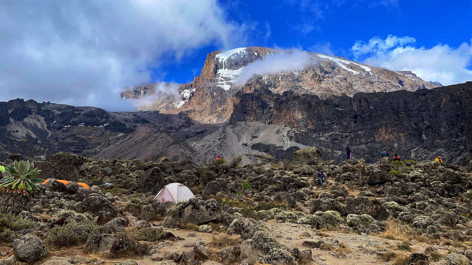 A tent sits among boulders. A tall peak rises behind it.