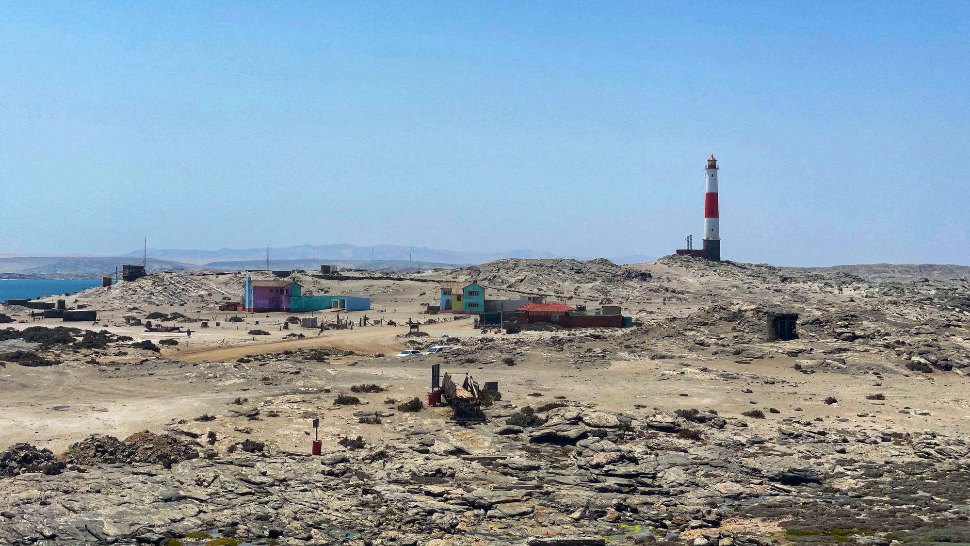 A candy cane striped lighthouse with a few colorful buildings on a rocky and barren landscape.