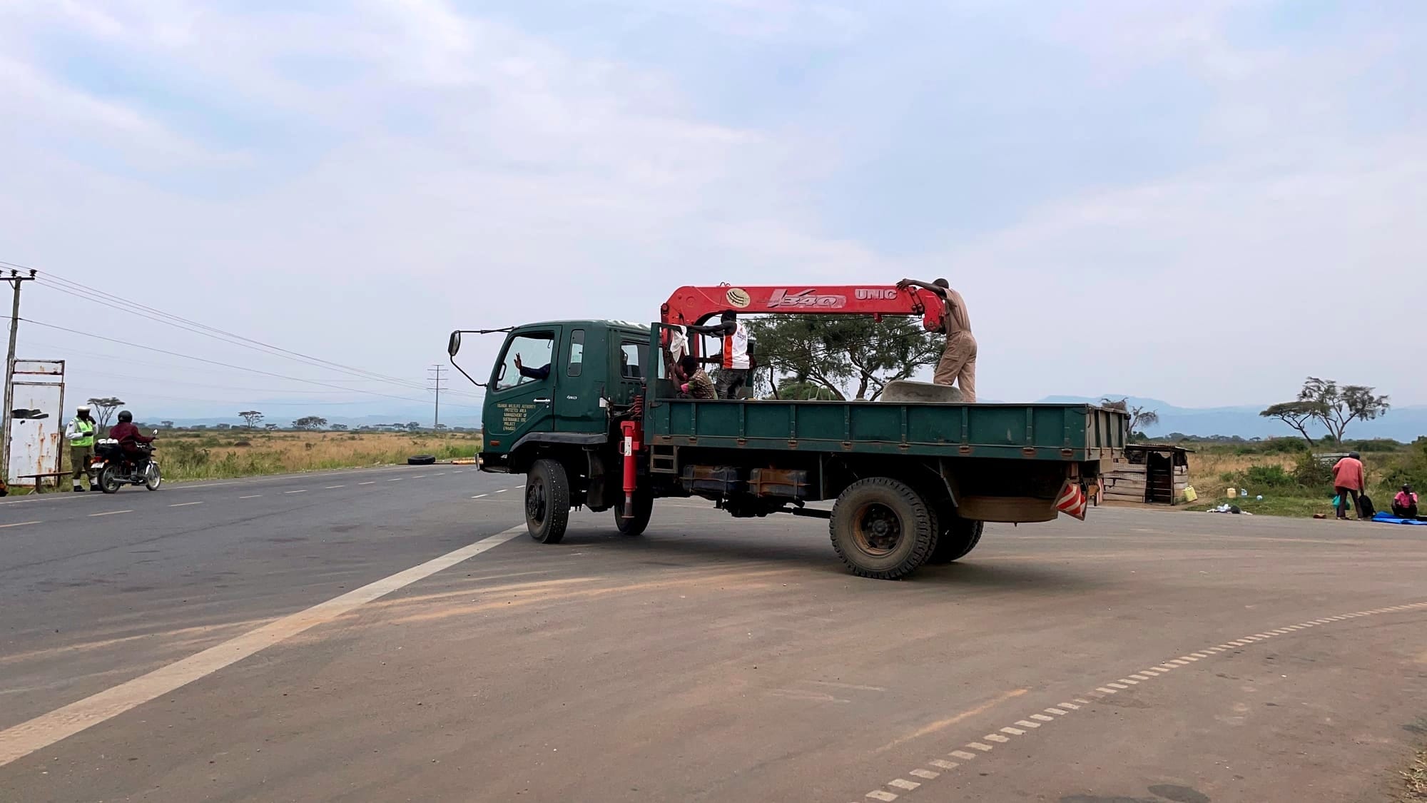 A green construction truck drives in the opposite direction with men standing in the back, and a waving hand out the window.