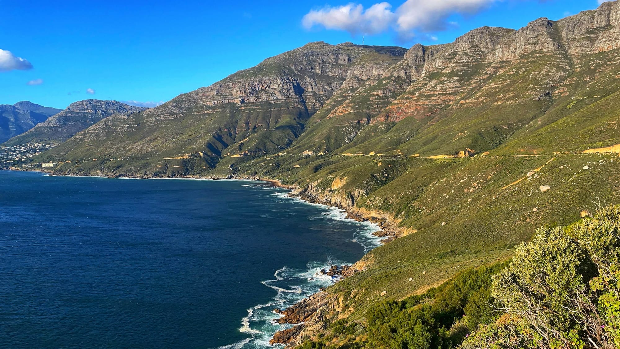 A road cuts through the side of a rocky mountain with low green vegetation that cascades into the ocean.