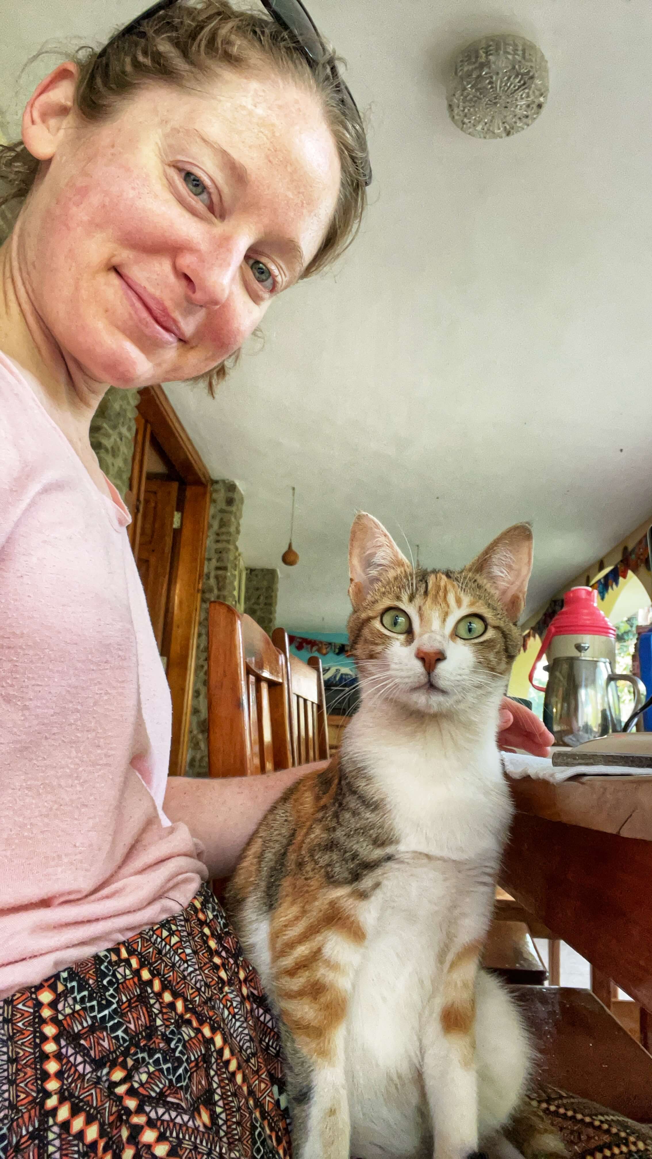 A woman sits at a wooden table with a calico cat on her lap. They both look at the camera.