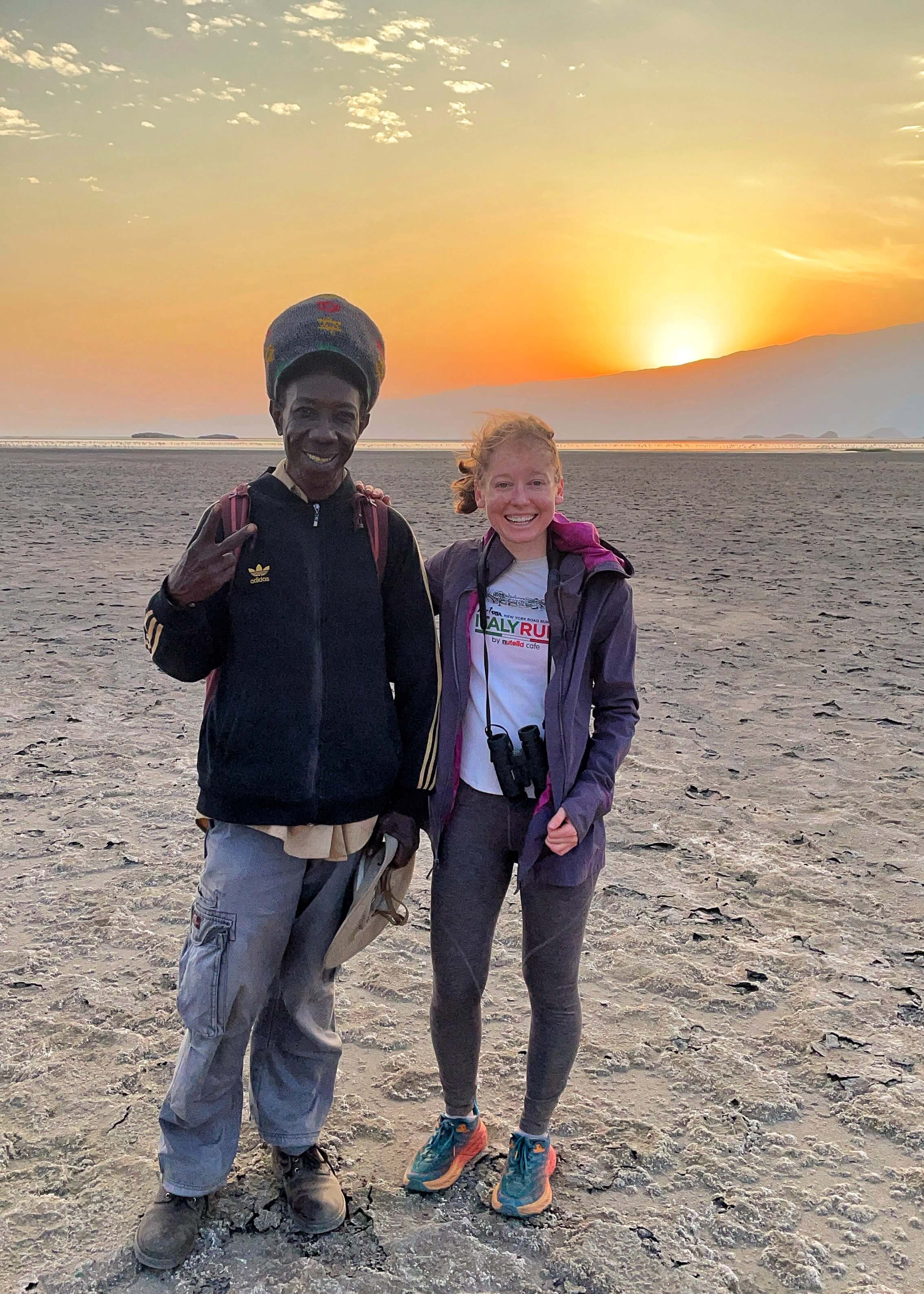 A white woman and an African man pose for a picture on a grey, dry salt bed as the sun rises over a hill behind them.