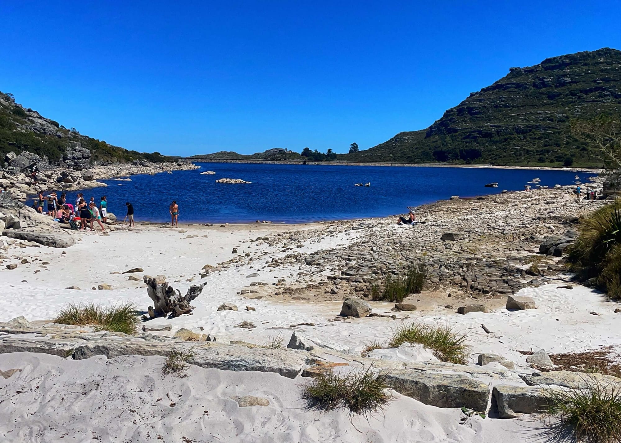 A blue lake and white sandy beach, with rocks and tall hills rising up around.