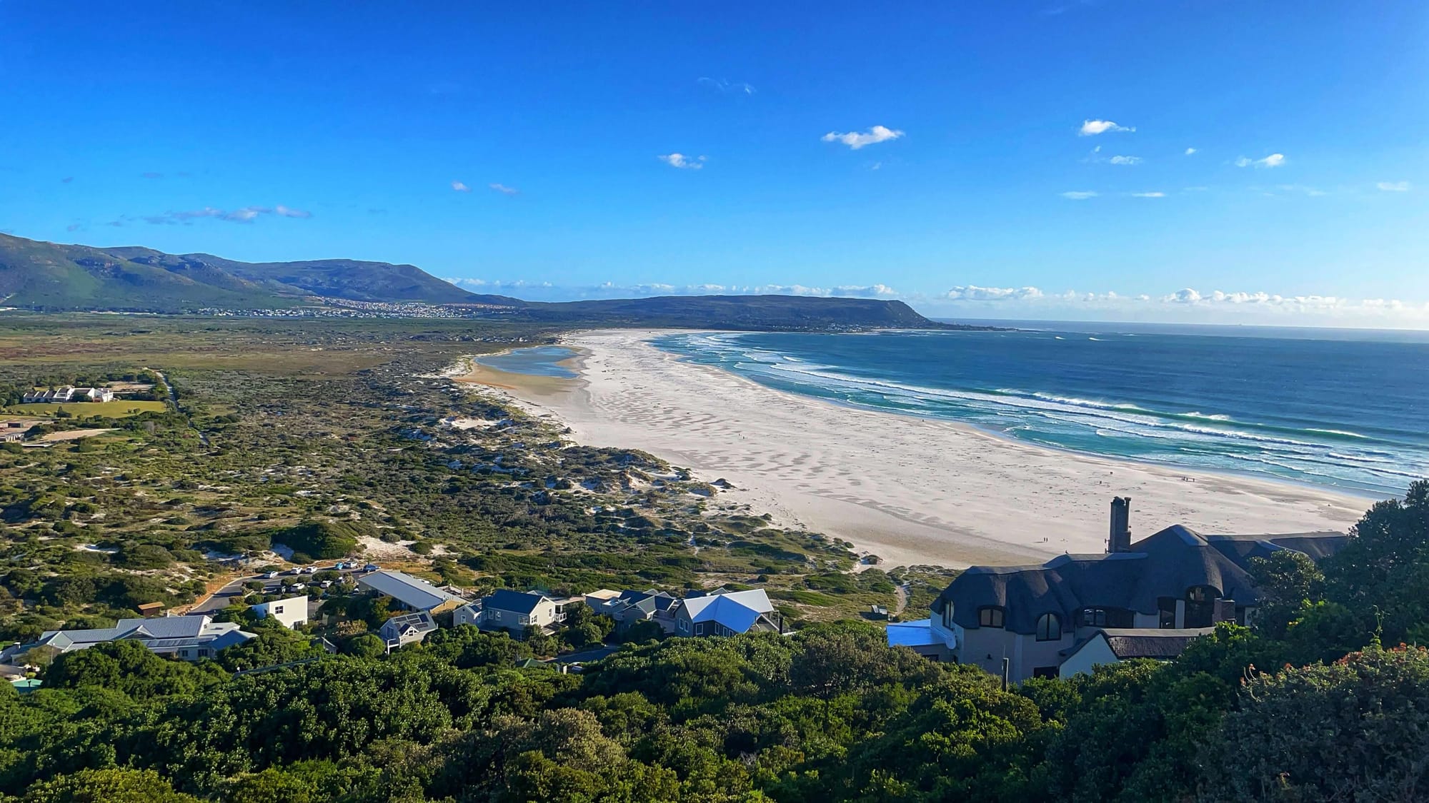 The view from above of houses along the cliff, and below, a long white sand beach and ocean at low-tide.