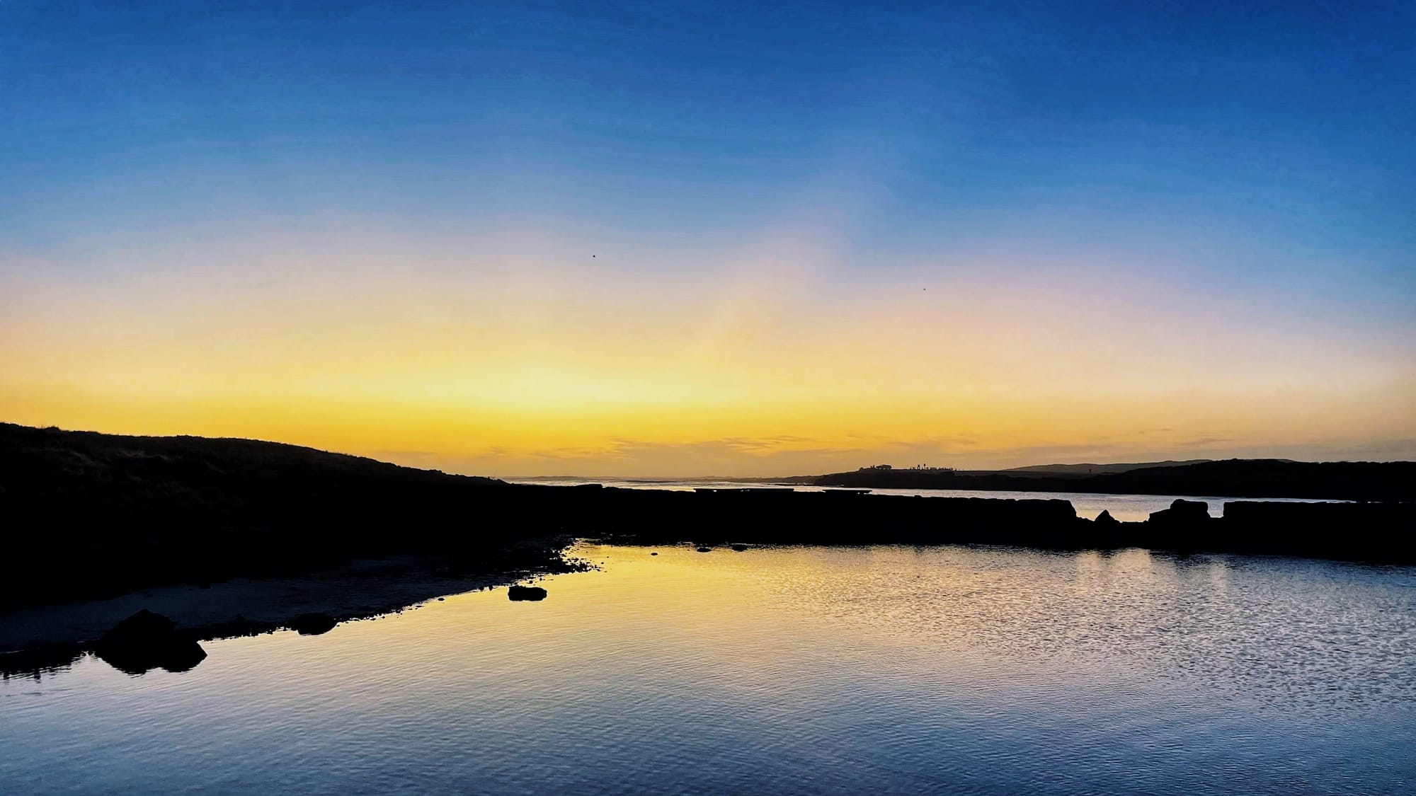 At dusk, a yellow glow on the horizon gives way to a deep blue sky. The glow reflects on calm waters.