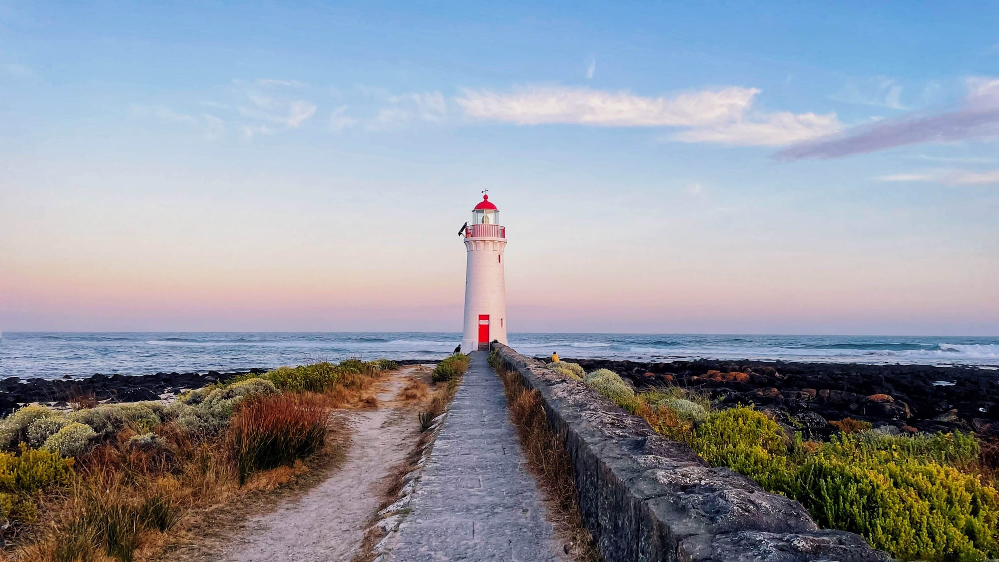 The sky is painted pastel blue, pink and faint yellow at sunset. A white lighthouse with a red door and roof carries a pink hue from the sun. It sits at the end of a pathway, surrounded by brush and black lava rock.