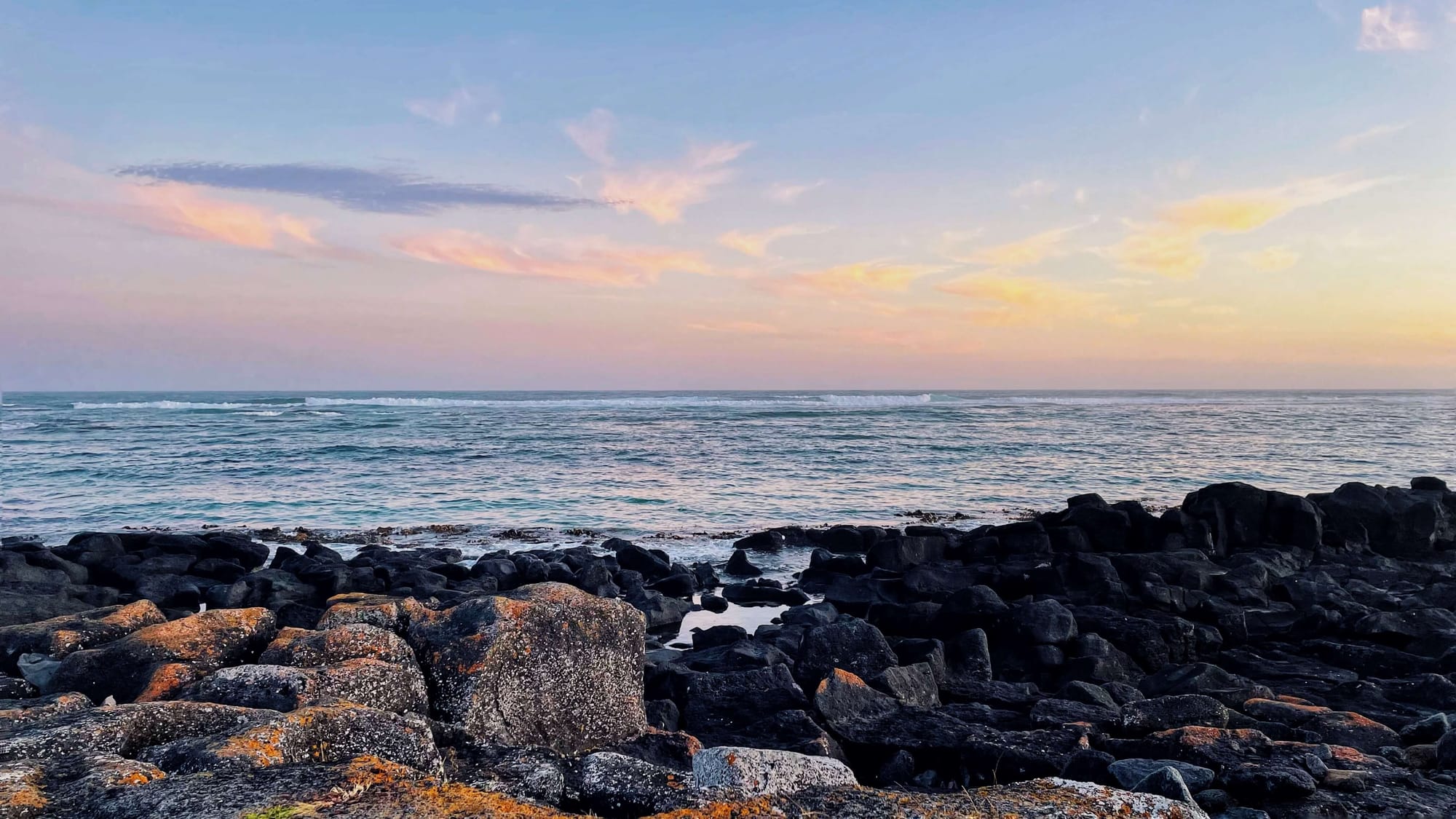 The sky is pink and it reflects onto the ocean. In the foreground are black lava boulders.