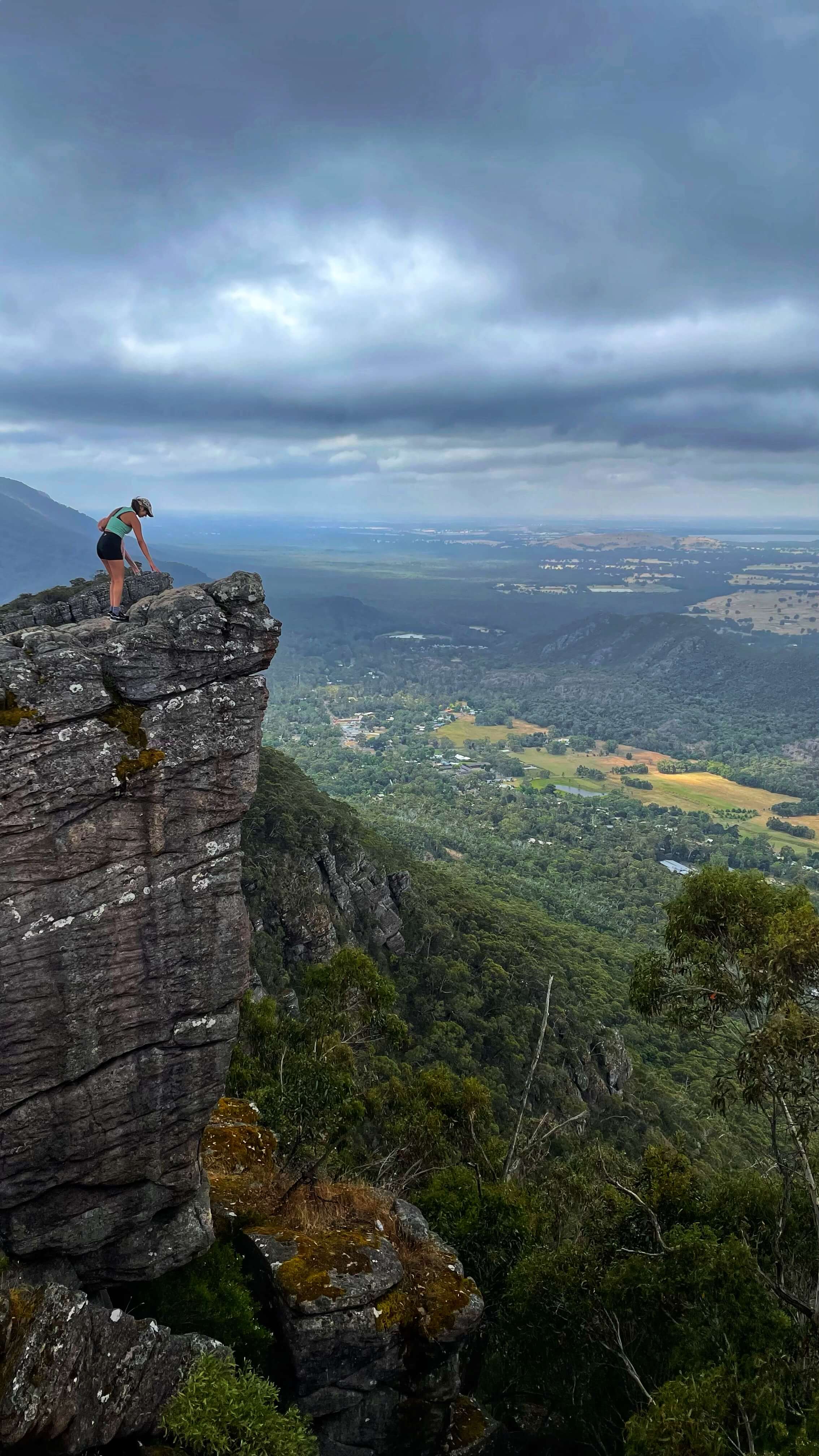 A woman stands at the edge of a lookout point on the steep sandstone cliffs. In the valley below is a town and green fields.