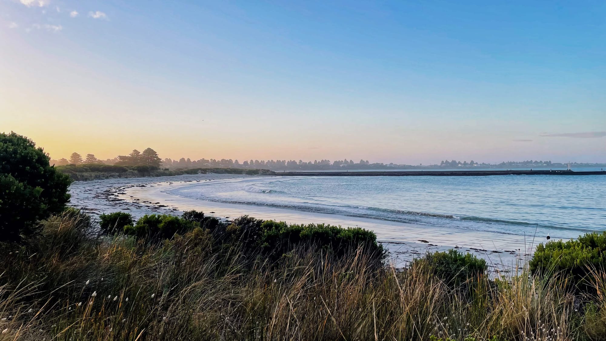 A c-shaped bay with white sand, and the view of a town in the far distance. The sky has a pastel hue, at sunset.