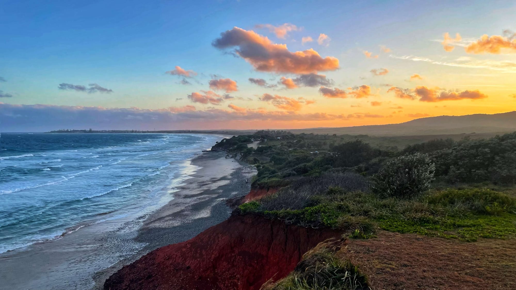 Red cliffs topped with green grasses cascading onto a white sand beach at sunset. The sky is painted blue, pink, orange and yellow.