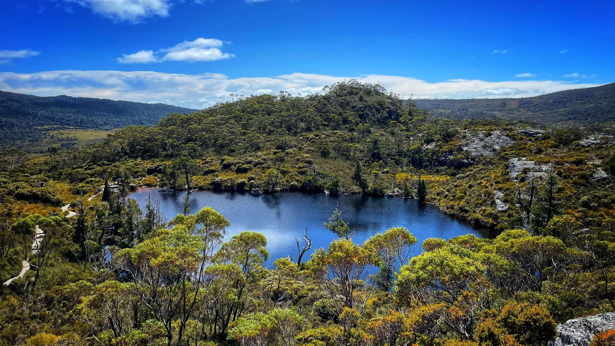 A deep blue alpine lake on a sunny day, surrounded by grey rocks and green trees and shrubs, with a boardwalk along the left side.