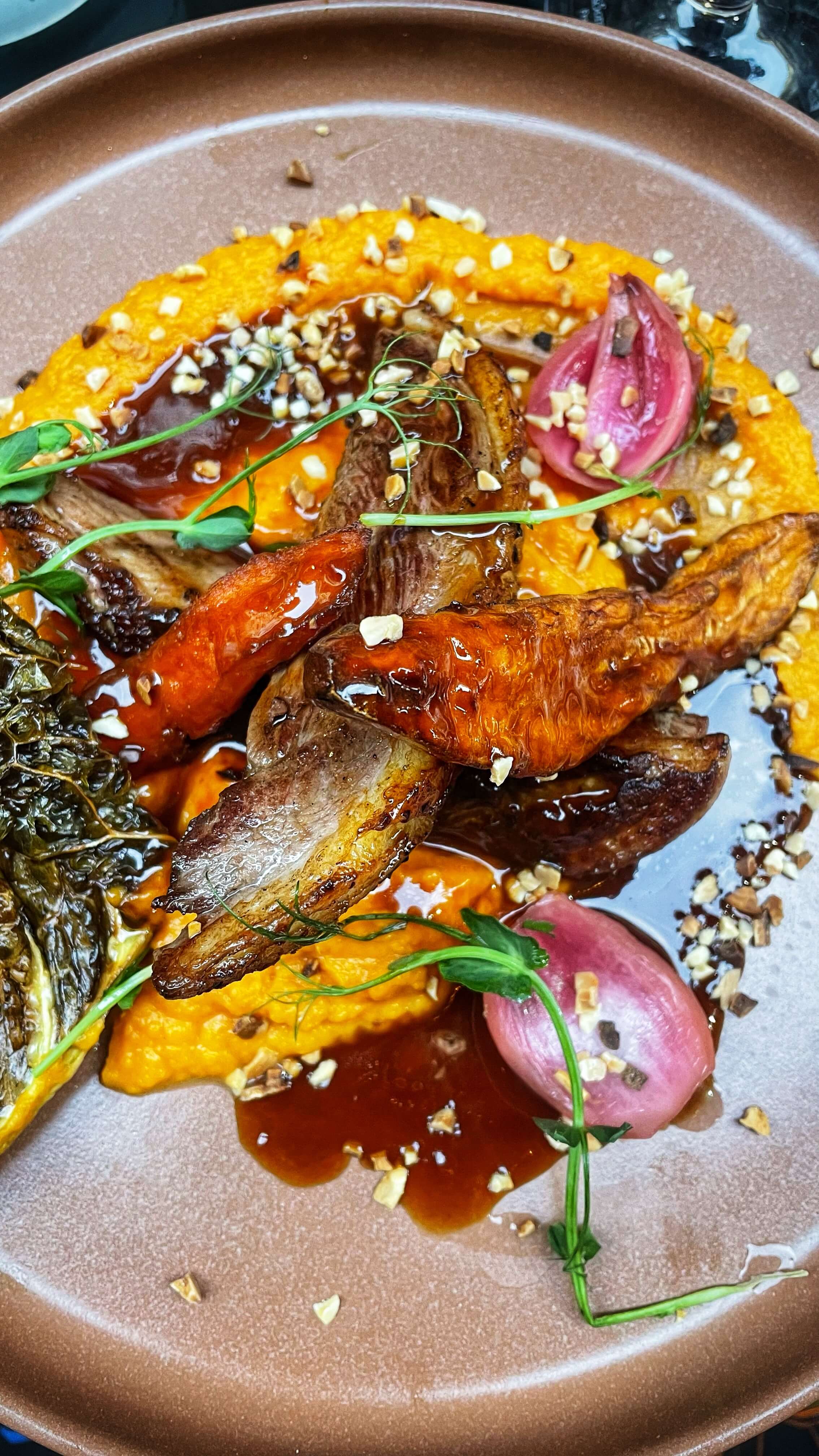 Yellow orange and green root vegetables on top of a pumpkin puree, with roasted duck, arranged on a plate like a work of art.