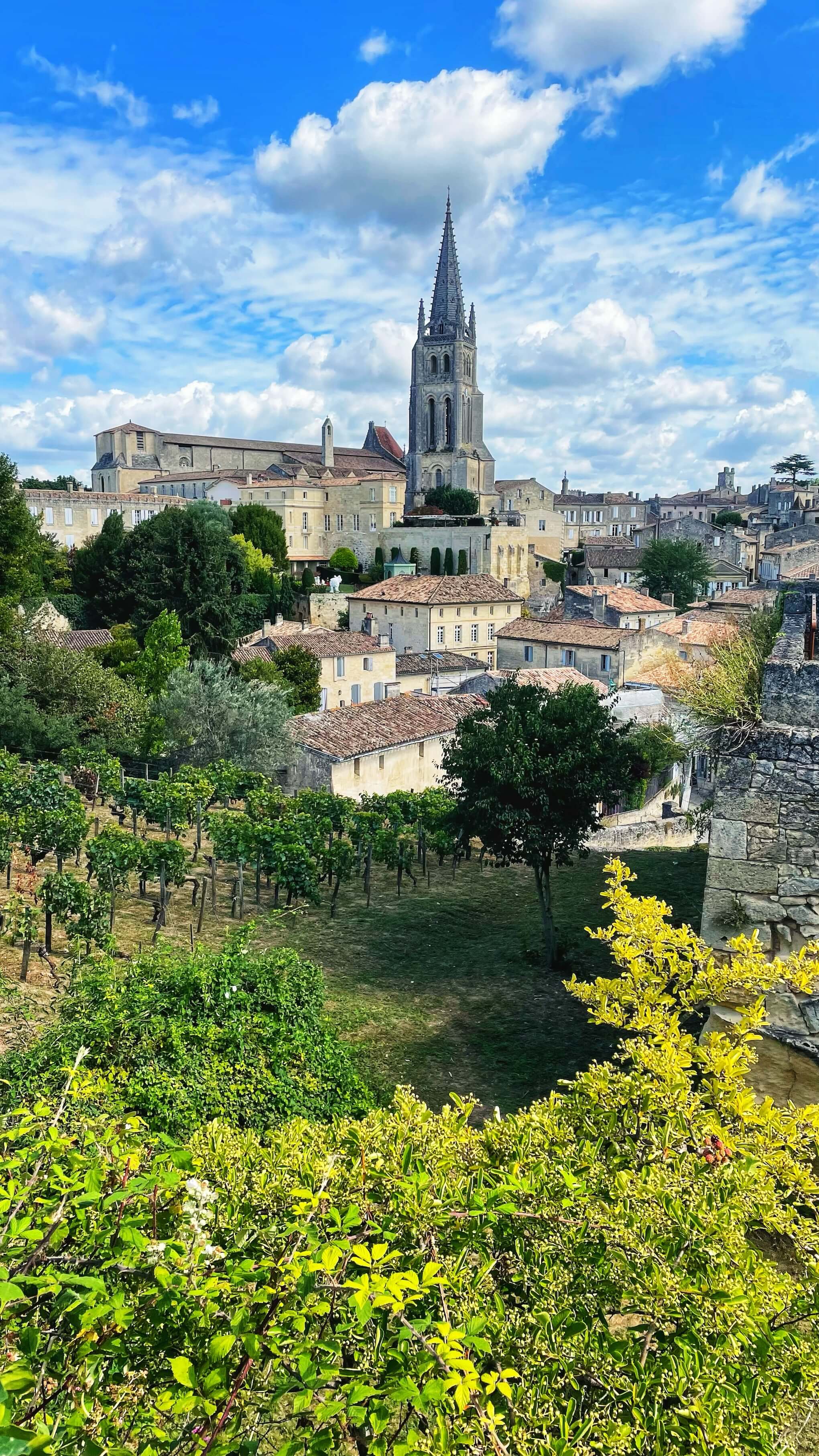 A view over Saint-Émilion, France, a compact village built with yellow and grey stones, with a vineyard in sight, and the tall steeple of the Église monolithe in the center of the village.