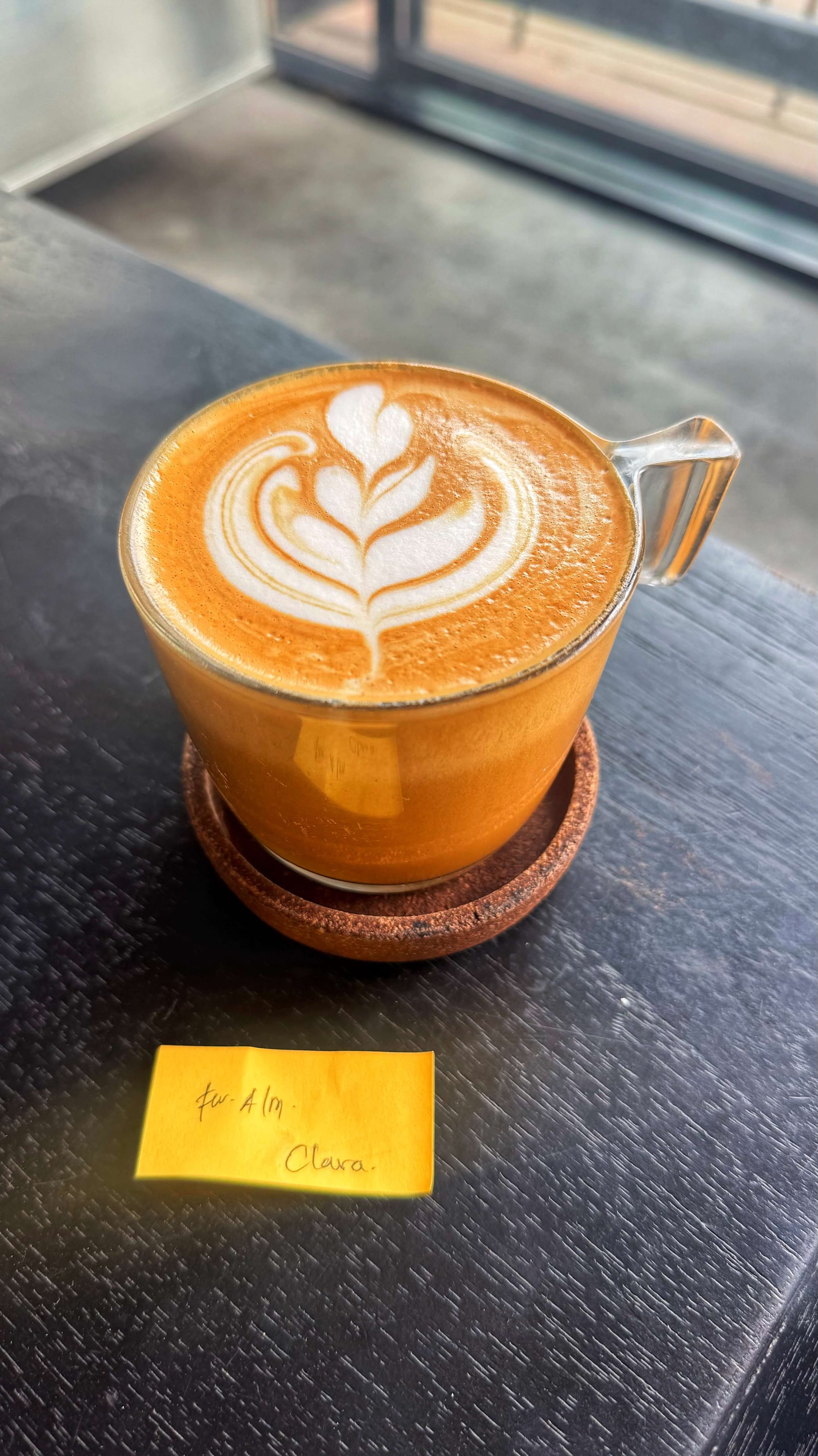 A flat white coffee with latte art and a sticky note that reads "Clara."