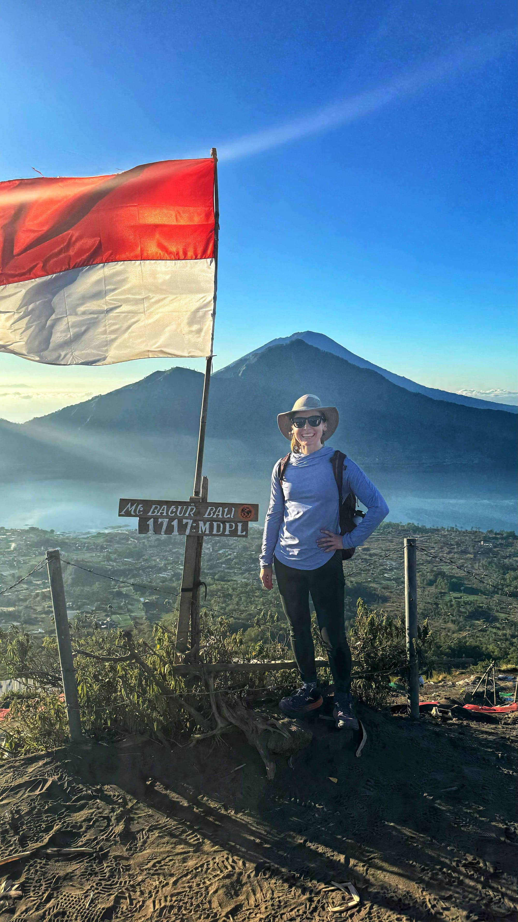 Clara Ritger poses next to the Indonesian flag at the summit of Mount Batur, overlooking a lake and opposing peak.