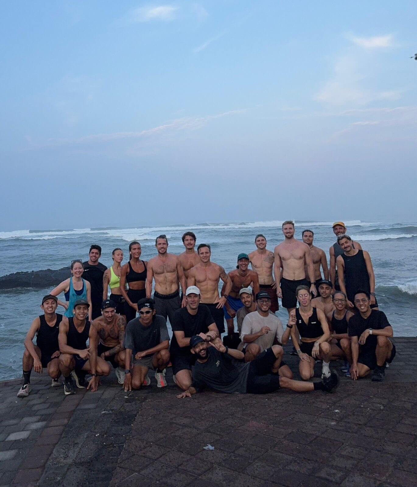 Twenty-five runners pose for a photo at sunrise in front of the beach.