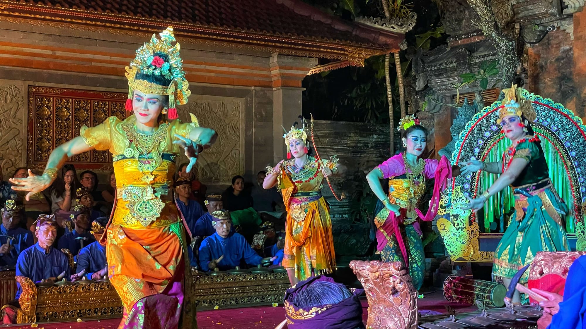 Four Balinese women dance in colorful costumes, makeup and head adornments, while seated men in blue play instruments.