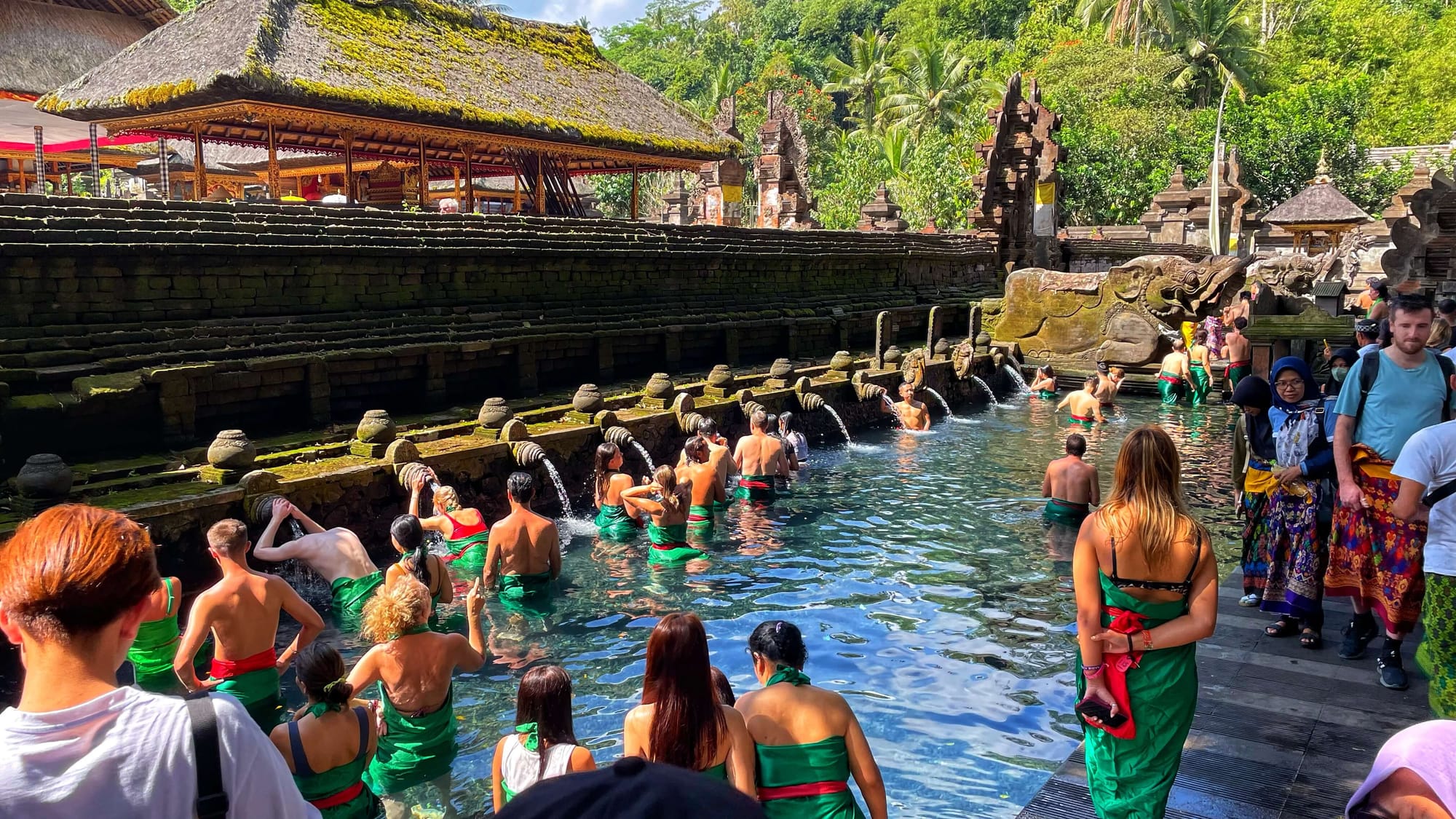 A long queue of tourists in green sarongs wait to enter a pool at a temple. 