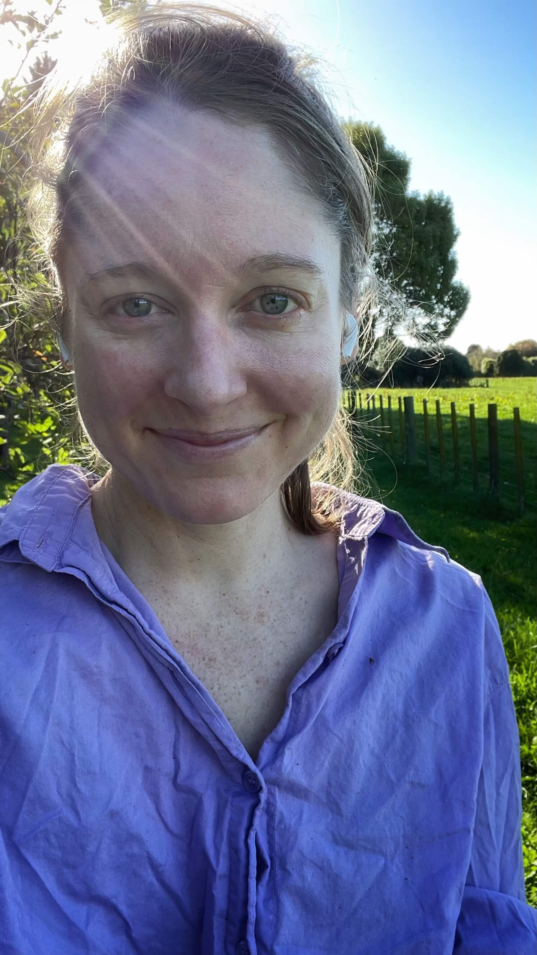 Clara Ritger smiles gently in a purple blouse in the midst of an orchard with the rays of the sun breaking over her head.
