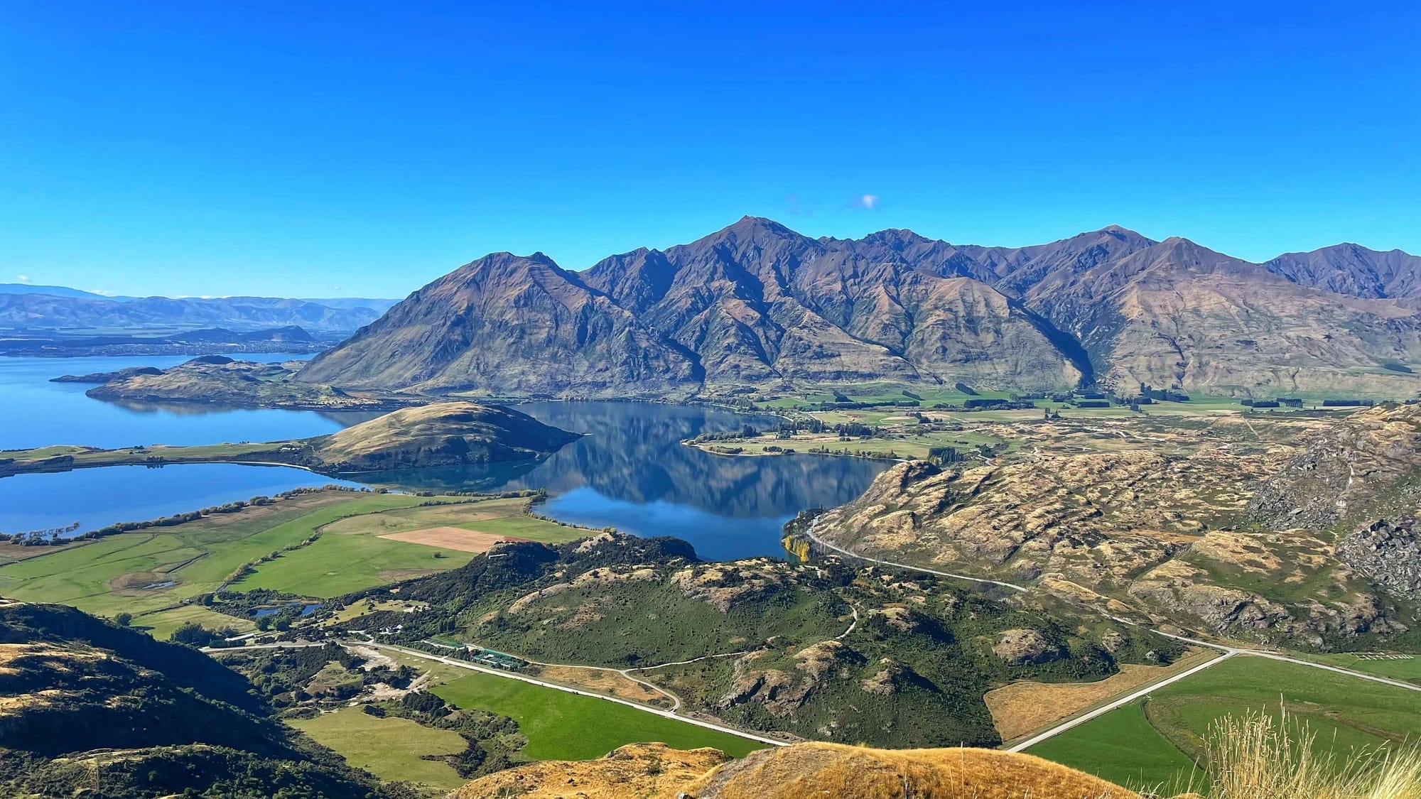A view over a lake and green pasture fields with rocky hills and mountains cut into the landscape.