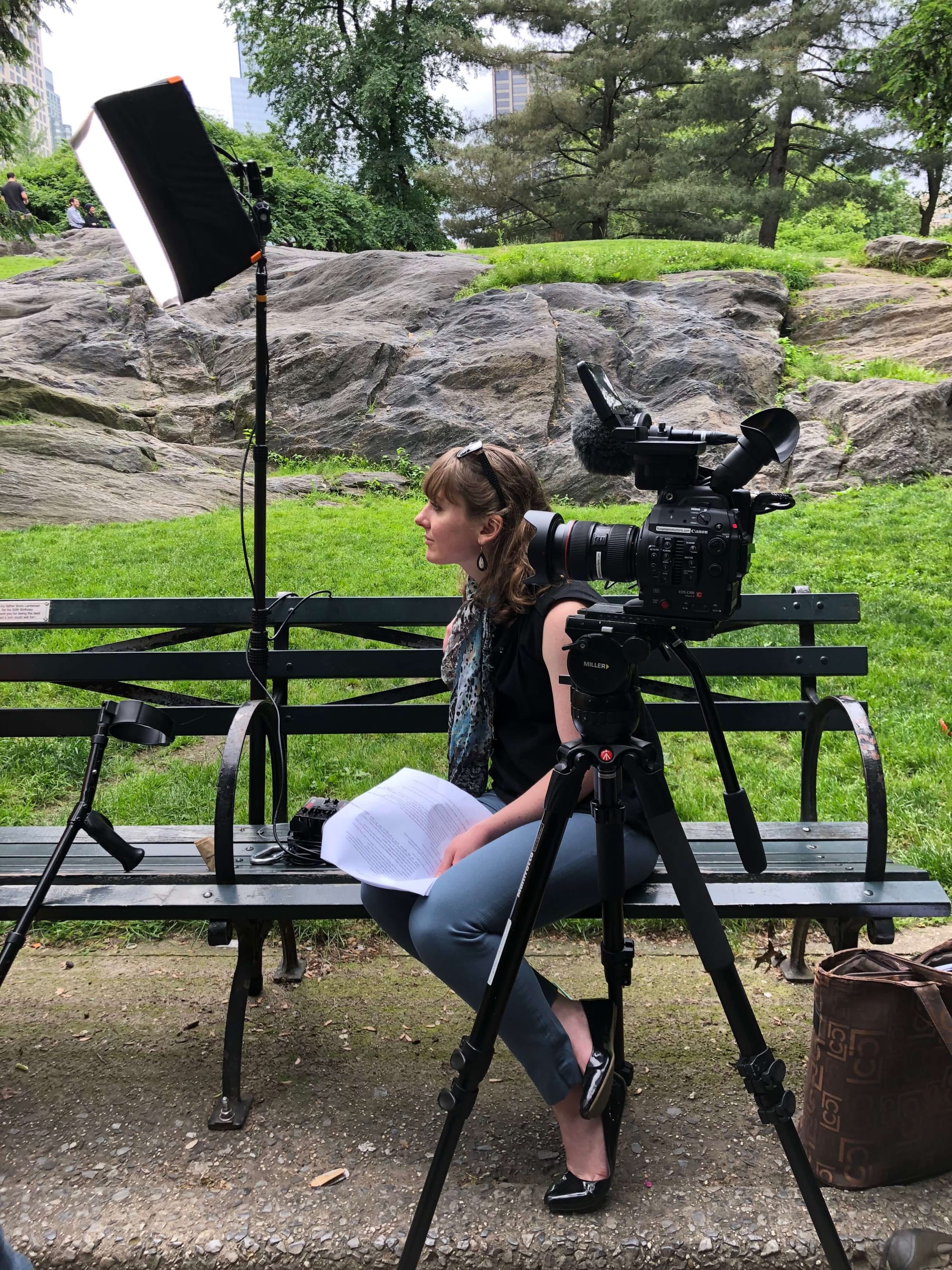 Clara Ritger sits on a park bench surrounded by a video camera and lights while she interviews.