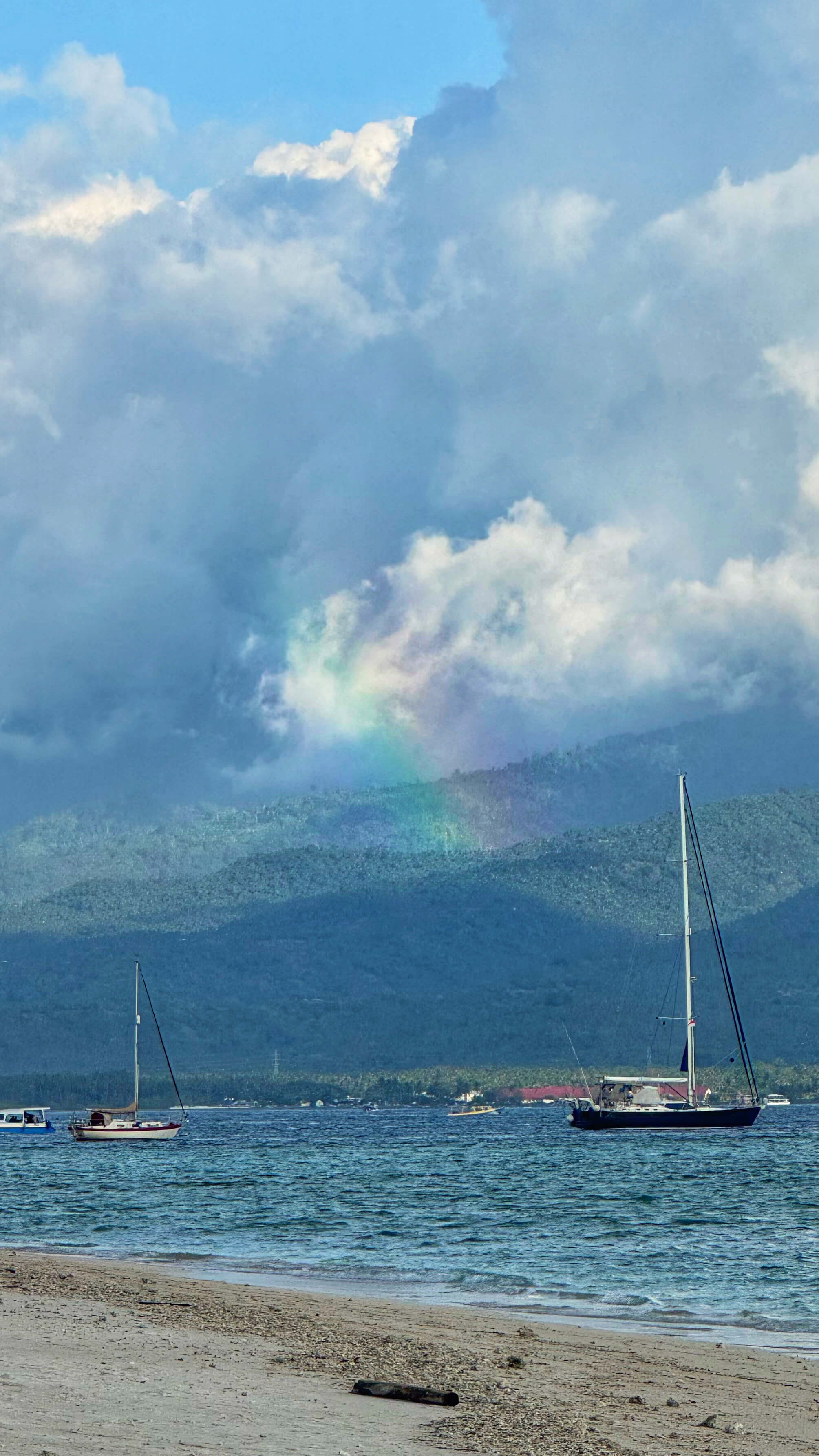 A rainbow over green mountains, with sailboats on the ocean in the foreground.