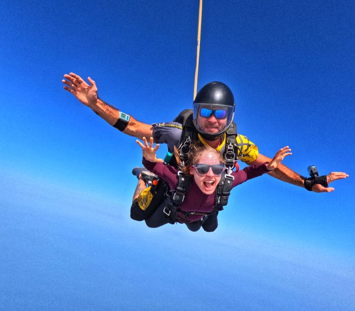 Clara Ritger and a male instructor tandem skydive against a blue sky with arms spread wide.