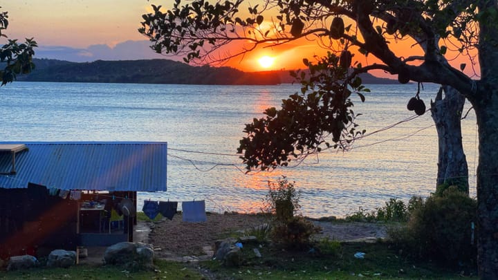 View from an island shore of an orange and pink sunset over another body of land in the lake.