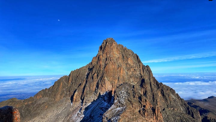 A triangular shaped, rocky mountain peak in a blue sky with a small moon behind.