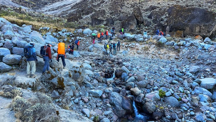A line of hikers is seen crossing a small waterfall surrounded by rocks.