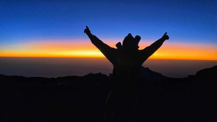 High in the sky, a silhouette raises its hands into the air in a triumphant "v" at sunrise.
