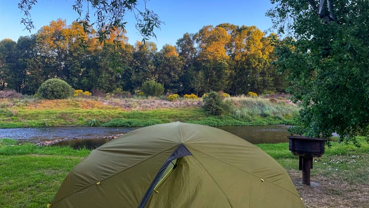 A green tent next to a river and an outdoor grill. The treetops are bathed in yellow , at sunset.