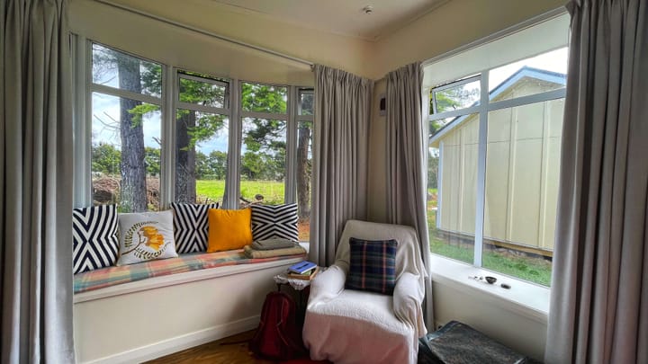 Two curtained windows in a cabin, with a loveseat and an armchair, looking out to trees and a green field. 