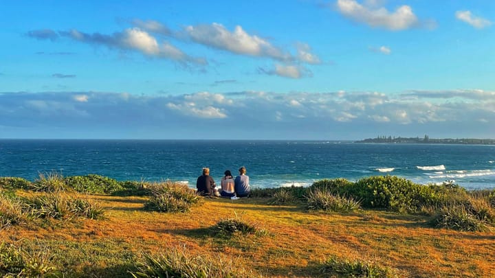Three girls look at the ocean, perched on golden yellow and orange grasses.