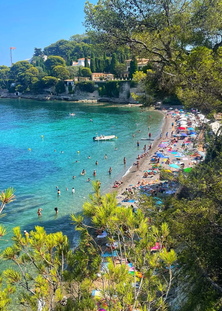 An overhead view of a beautiful blue beach in the Mediterranean, littered with colorful umbrellas and towels and people. 