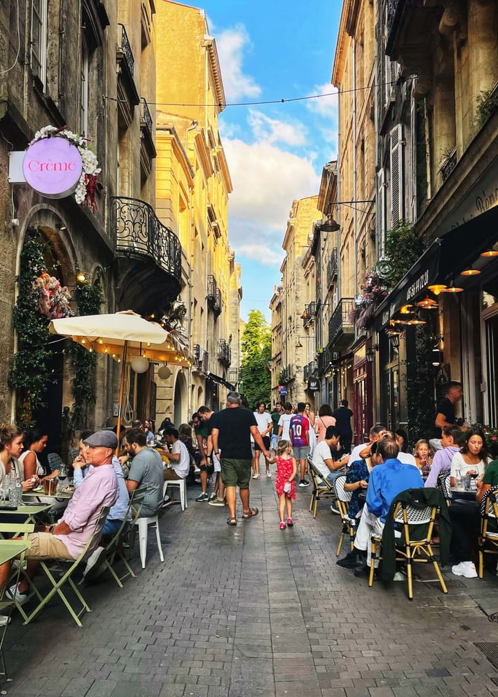 A dad and daughter walk hand-in-hand down a cobblestone alley lined with outdoor dining tables and filled with people.