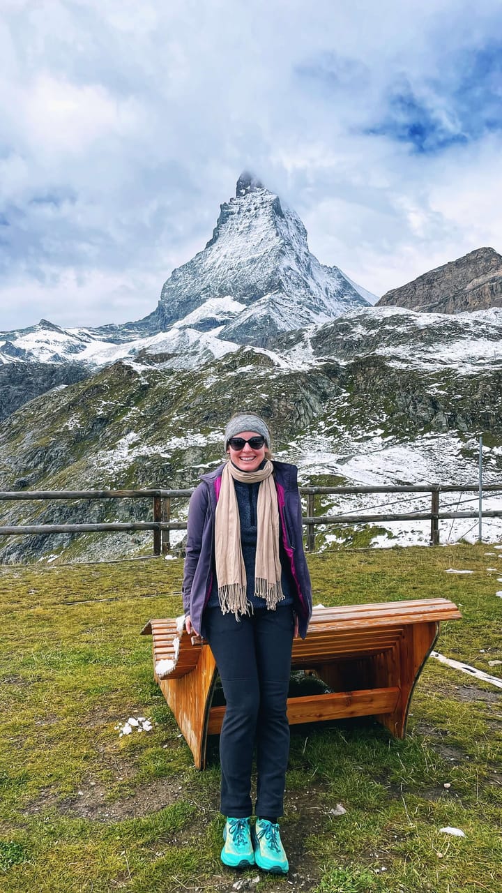 A woman bundled up in warm clothes poses in front of a uniquely shaped mountain peak that looks like a witch hat.
