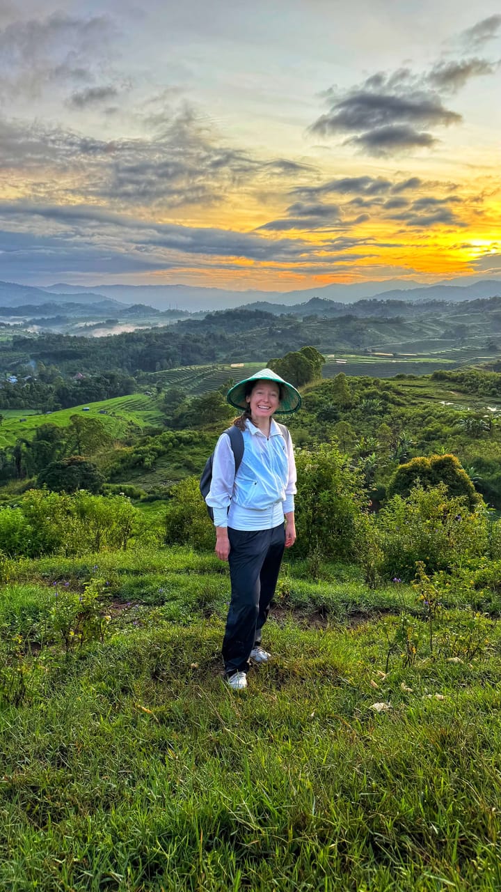 A woman smiles at the camera from the top of a green hill overlooking rice fields at s